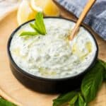 Small black dish sitting on wooden board of Cucumber and Yoghurt Dip, with a spoon resting in bowl.