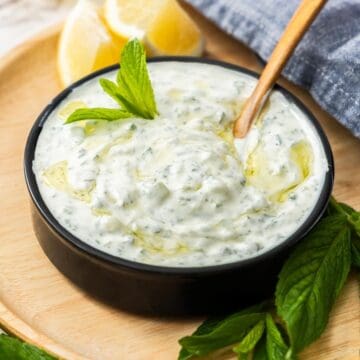 Small black dish sitting on wooden board of Cucumber and Yoghurt Dip, with a spoon resting in bowl.