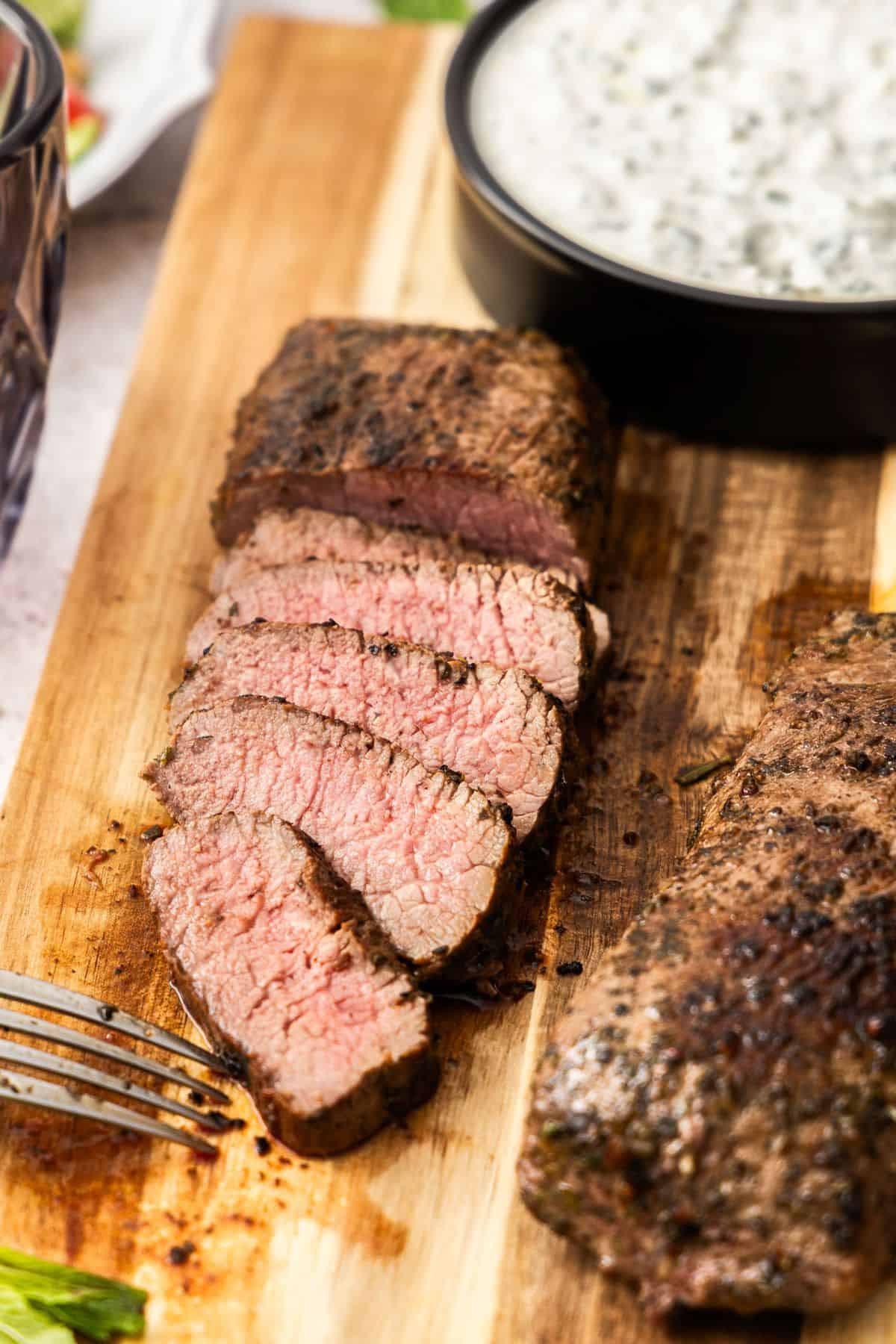 Wooden board, with dish of yoghurt sauce and two lamb backstraps, one slice into pieces.