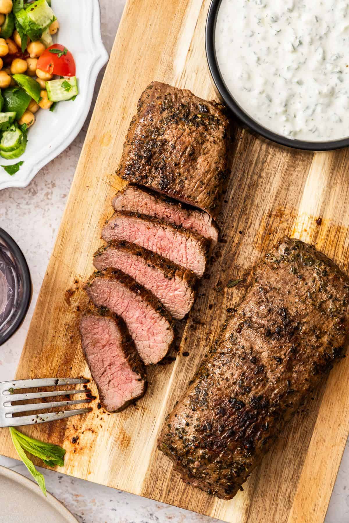 Wooden board, with dish of yoghurt sauce and two lamb backstraps, one slice into pieces.