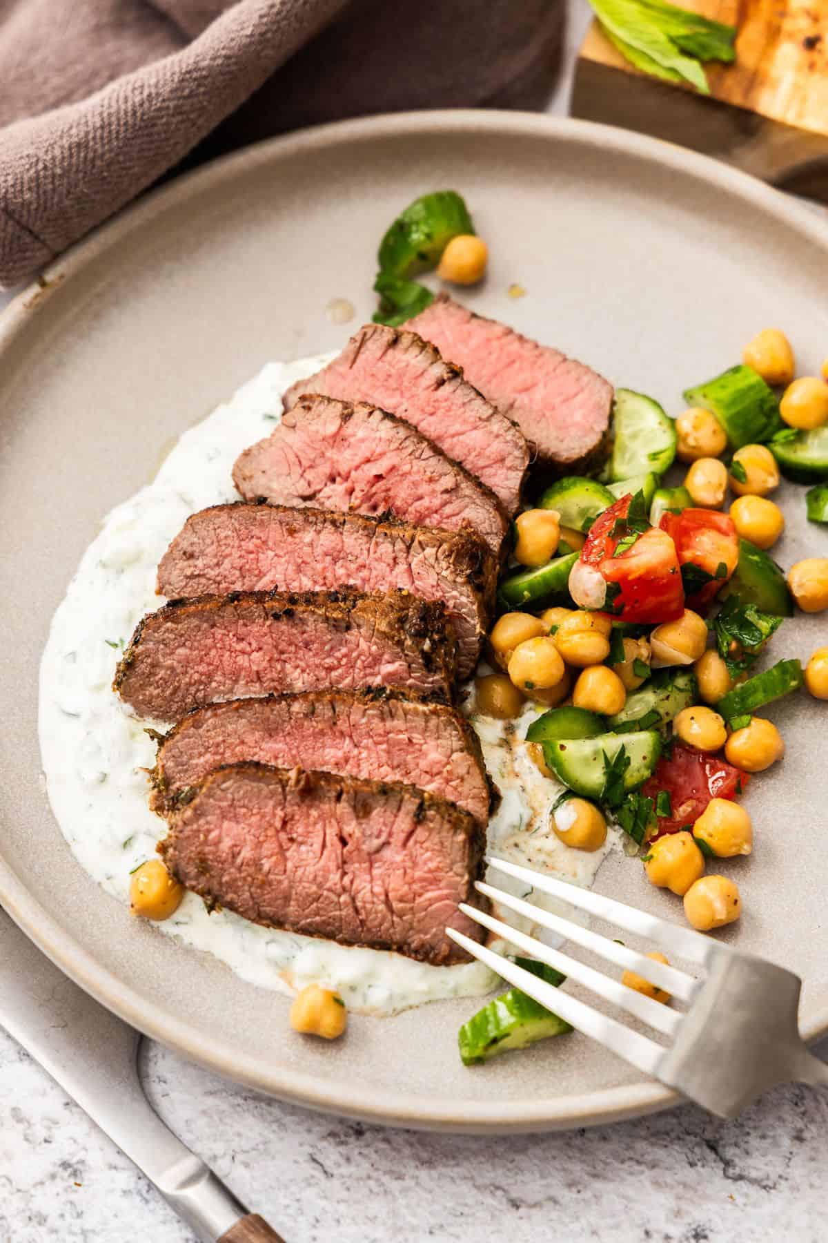 Round grey plate with a serving of sliced Lamb Backstrap, sitting on yoghurt sauce, with a chickpea cucumber side salad.