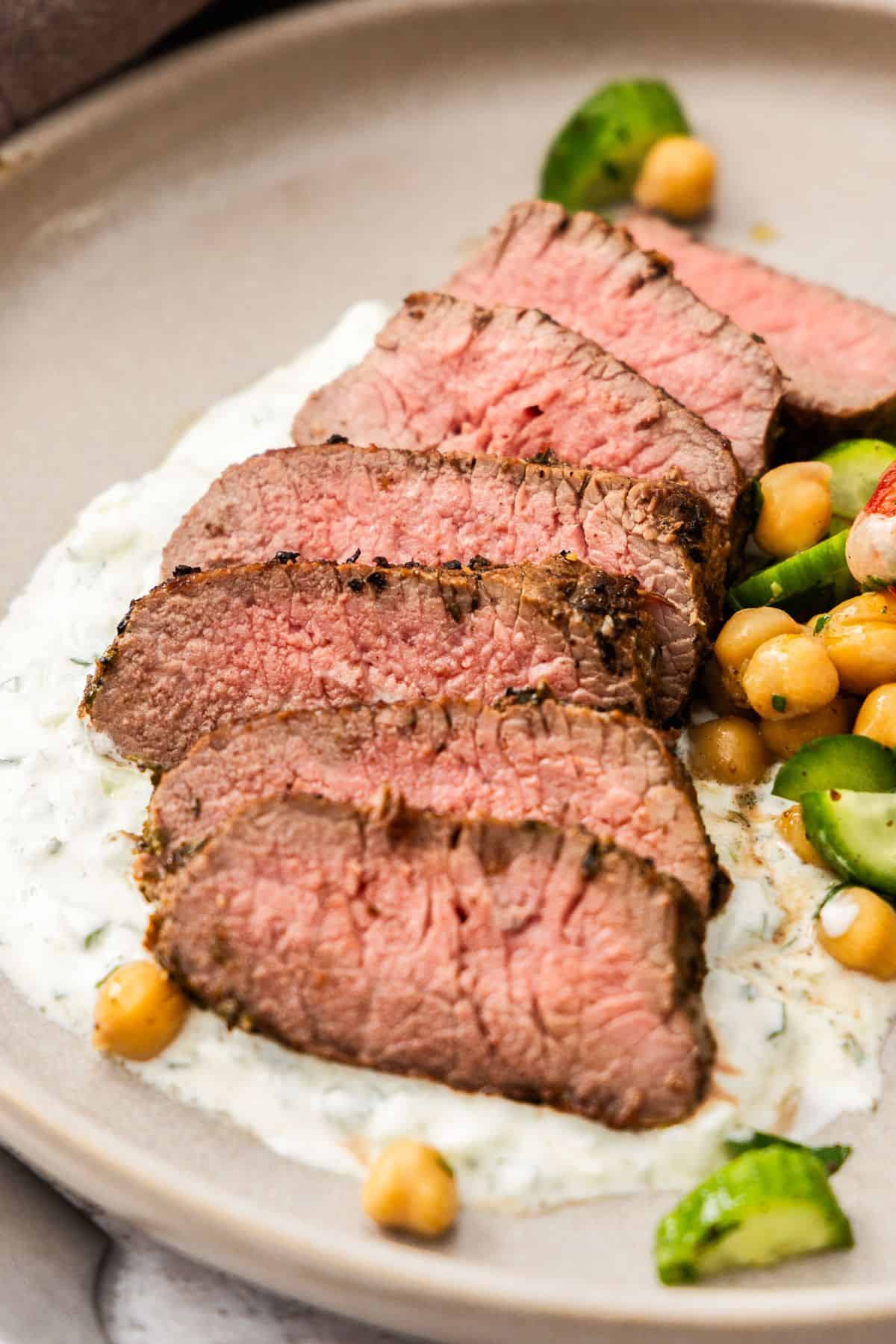 Round grey plate with a serving of sliced Lamb Backstrap, sitting on yoghurt sauce, with a chickpea cucumber side salad.