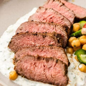 Round grey plate with a serving of sliced Lamb Backstrap, sitting on yoghurt sauce, with a chickpea cucumber side salad.