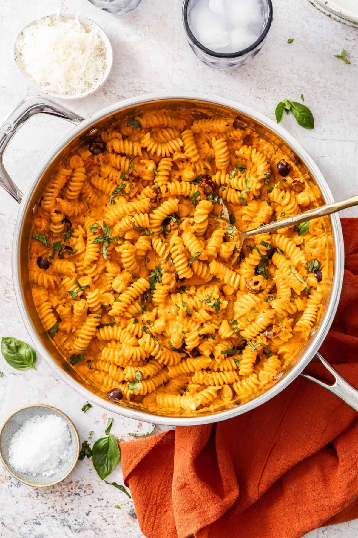 Large frying pan of Creamy Red Pepper Pasta, sitting on a red cloth, with a spoon resting in the pan.