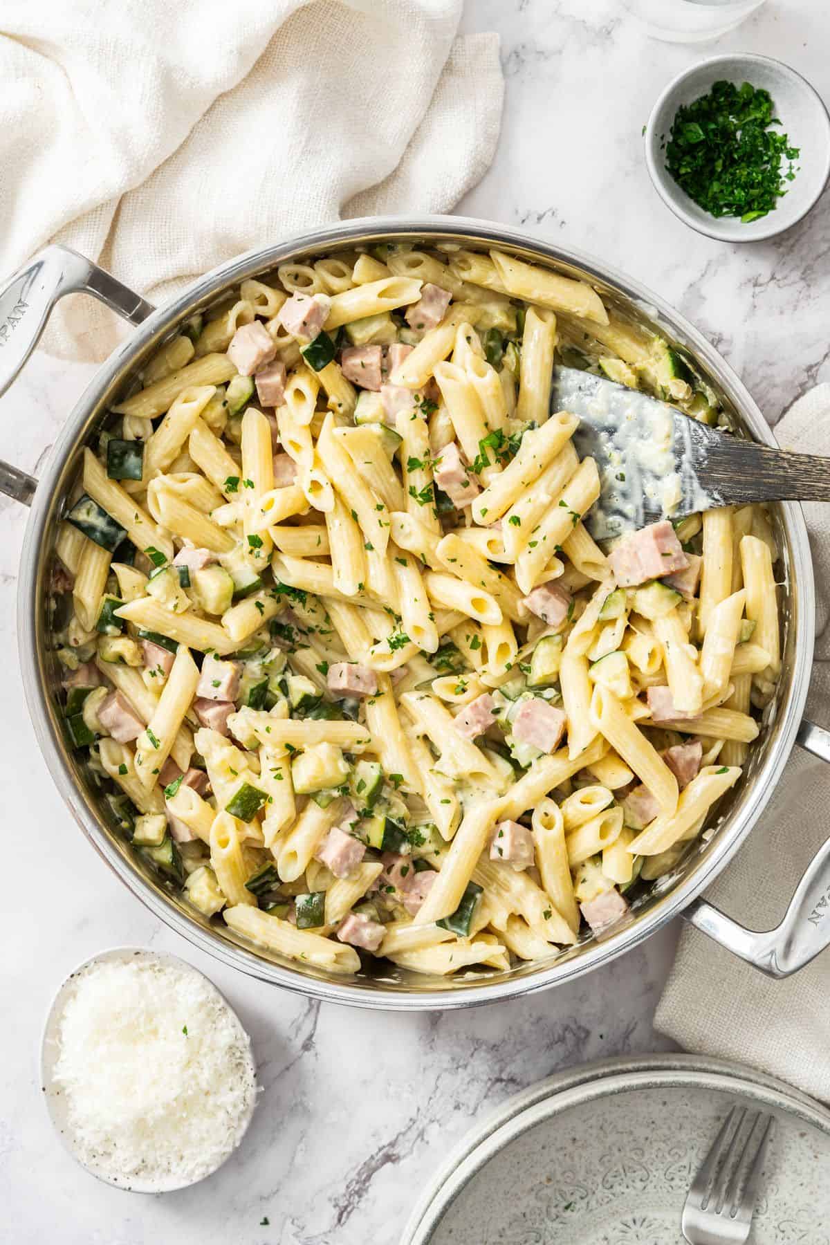 Large frying pan with Creamy Zucchini and Ham Pasta, with a wooden spoon resting in the pan.