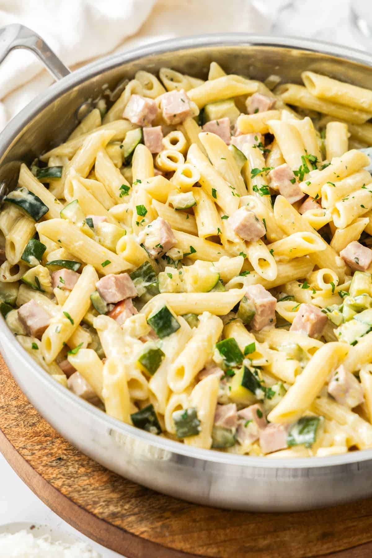 Large frying pan with Creamy Zucchini and Ham Pasta, sitting on a round wooden board.