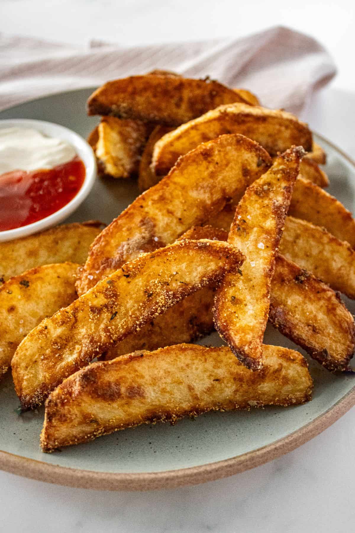 Round grey plate of Crispy Baked Potato Wedges, with a small dish of sour cream and sweet chilli sauce on the side.