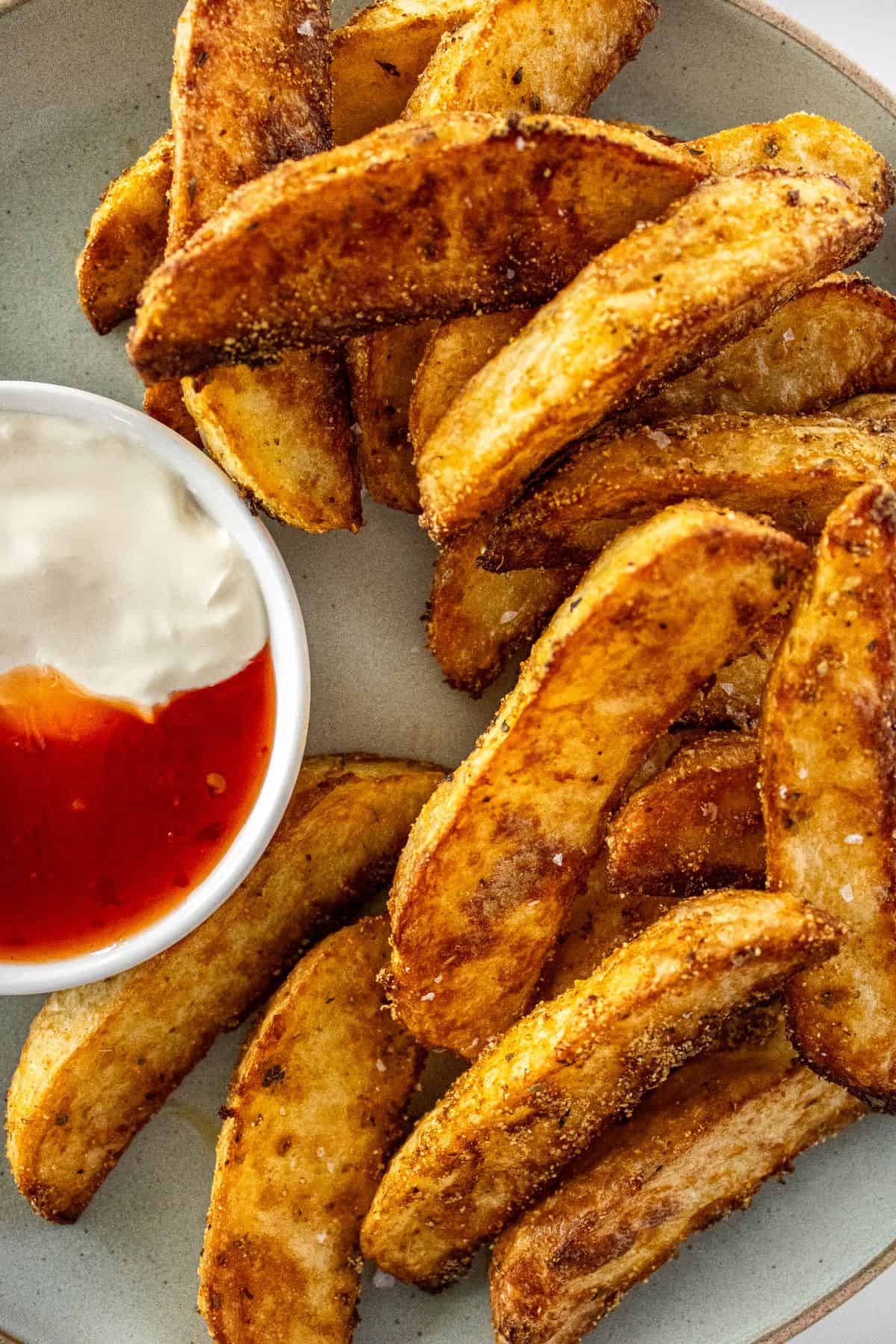 Round grey plate of Crispy Baked Potato Wedges, with a small dish of sour cream and sweet chilli sauce on the side.