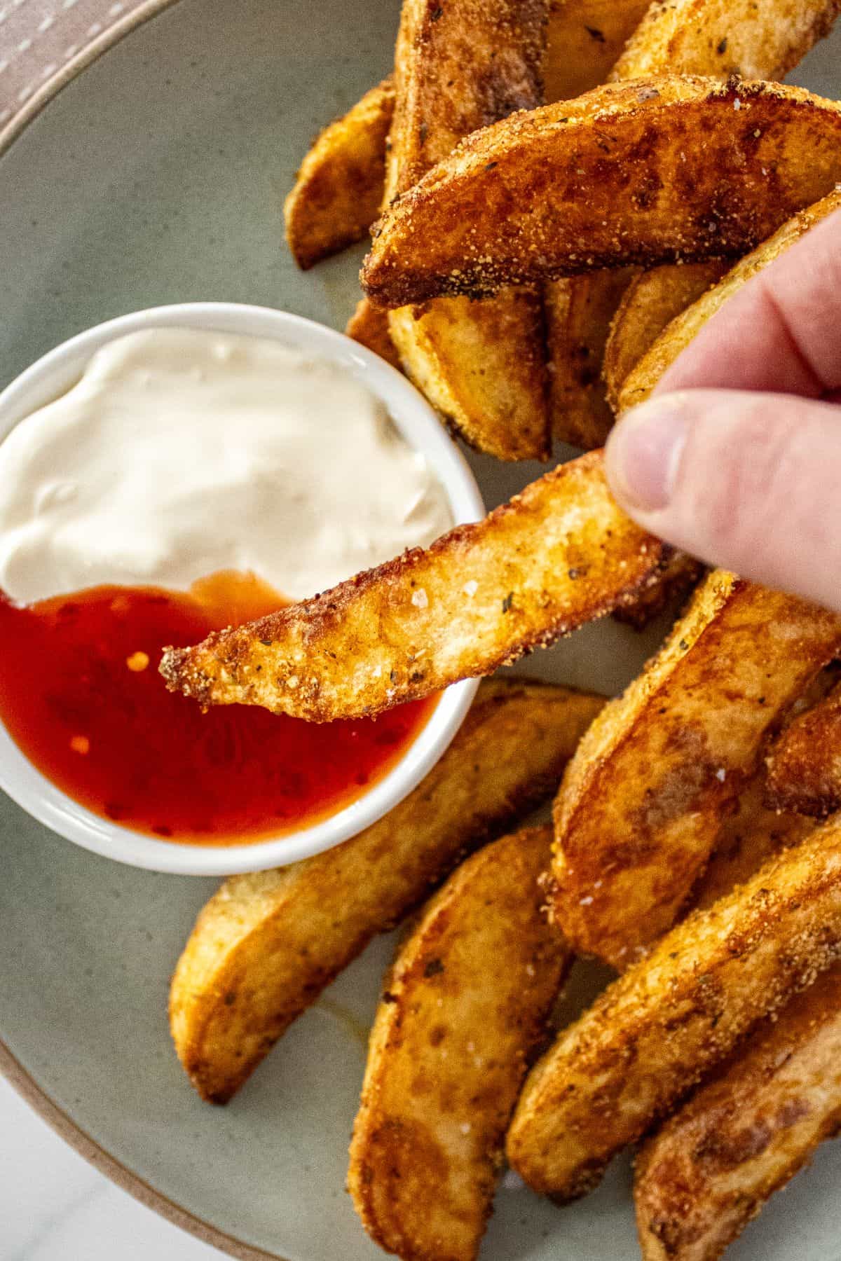Round plate of Crispy Baked Potato Wedges, with a hand dipping a wedge into a small dish of sour cream and sweet chilli sauce.