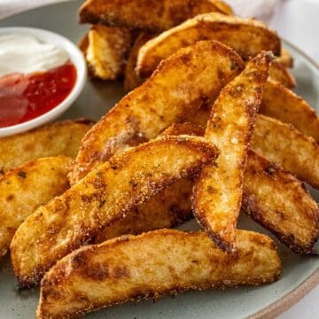 Round grey plate of Crispy Baked Potato Wedges, with a small dish of sour cream and sweet chilli sauce on the side.