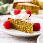 Piece of Pistachio Cake on a small white plate, with the cake topped with whipped cream and fresh raspberries.