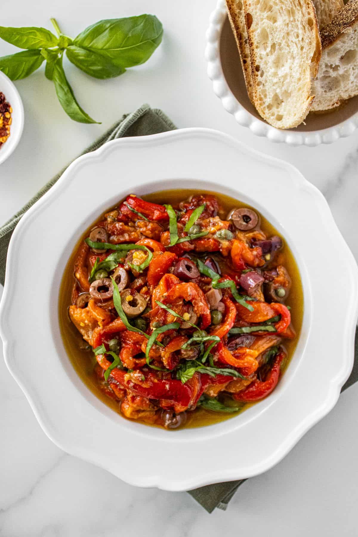 Round white bowl of Marinated Capsicums on a green cloth, with some basil, chilli and bread around the edge.