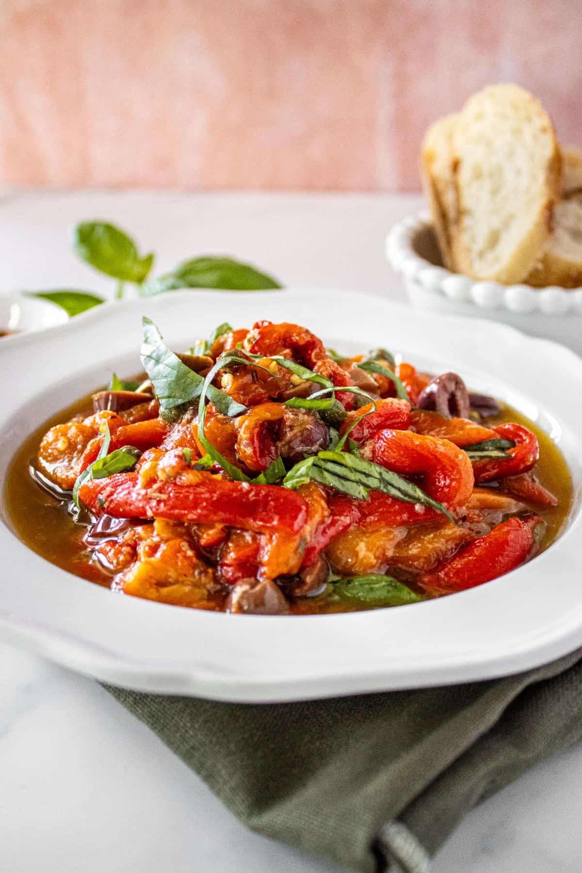 Round white bowl of Marinated Capsicums on a green cloth, with some basil, chilli and bread around the edge.