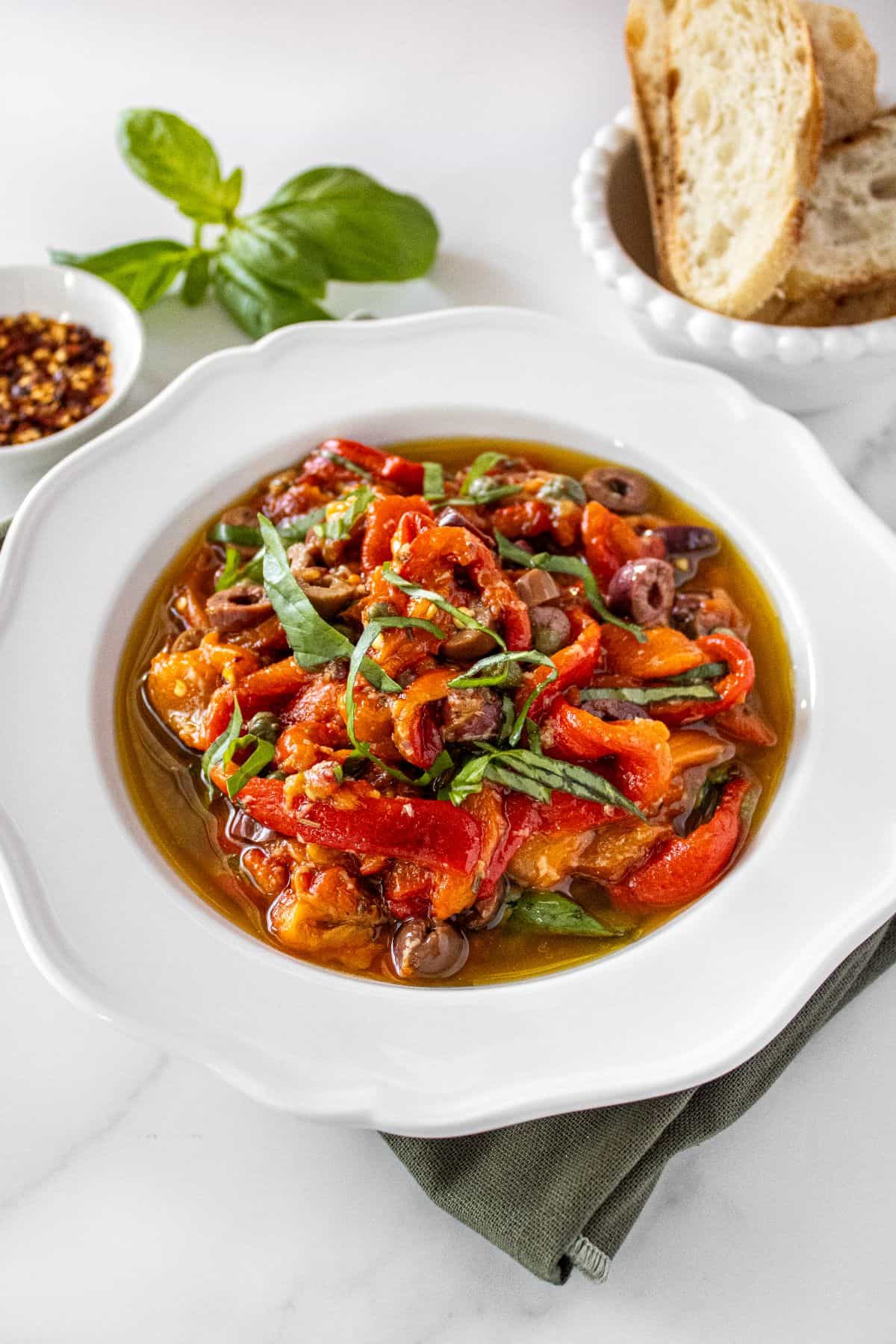 Round white bowl of Marinated Capsicums on a green cloth, with some basil, chilli and bread around the edge.