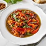 Round white bowl of Marinated Capsicums on a green cloth, with some basil, chilli and bread around the edge.