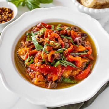 Round white bowl of Marinated Capsicums on a green cloth, with some basil, chilli and bread around the edge.