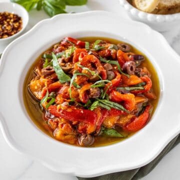 Round white bowl of Marinated Capsicums on a green cloth, with some basil, chilli and bread around the edge.