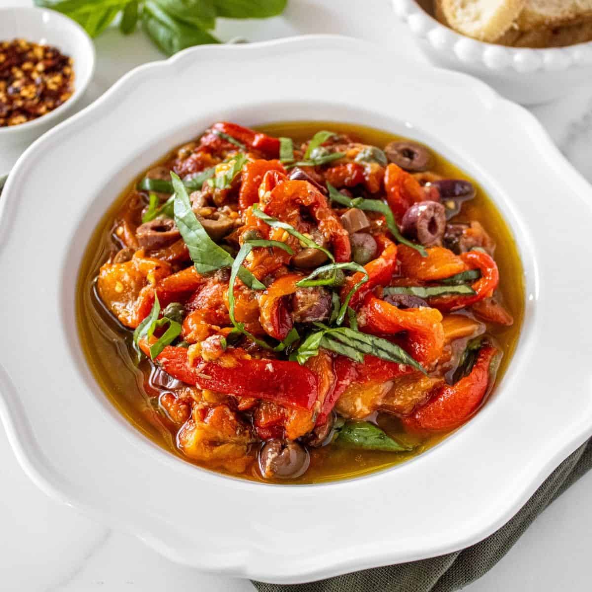 Round white bowl of Marinated Capsicums on a green cloth, with some basil, chilli and bread around the edge.