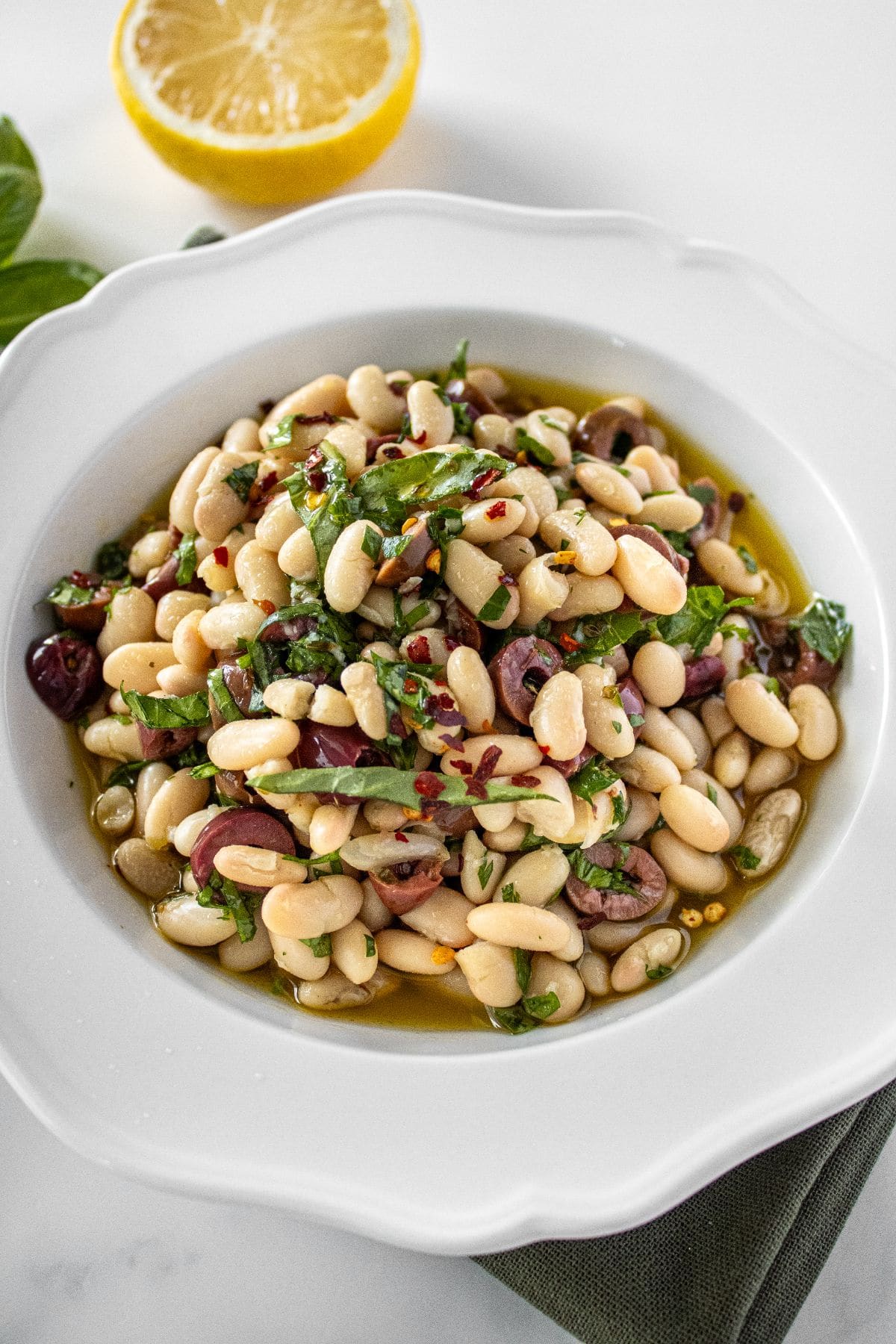 Round white bowl of Marinated White Beans, sitting on a green cloth, with some basil and lemon on the edge.