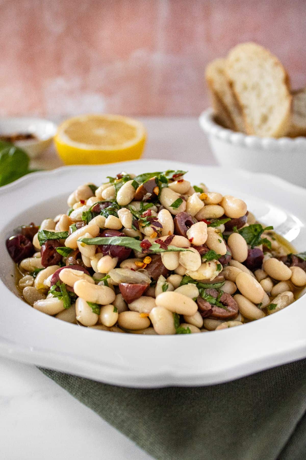 Round white bowl of Marinated White Beans, sitting on a green cloth, with some basil and lemon on the edge.
