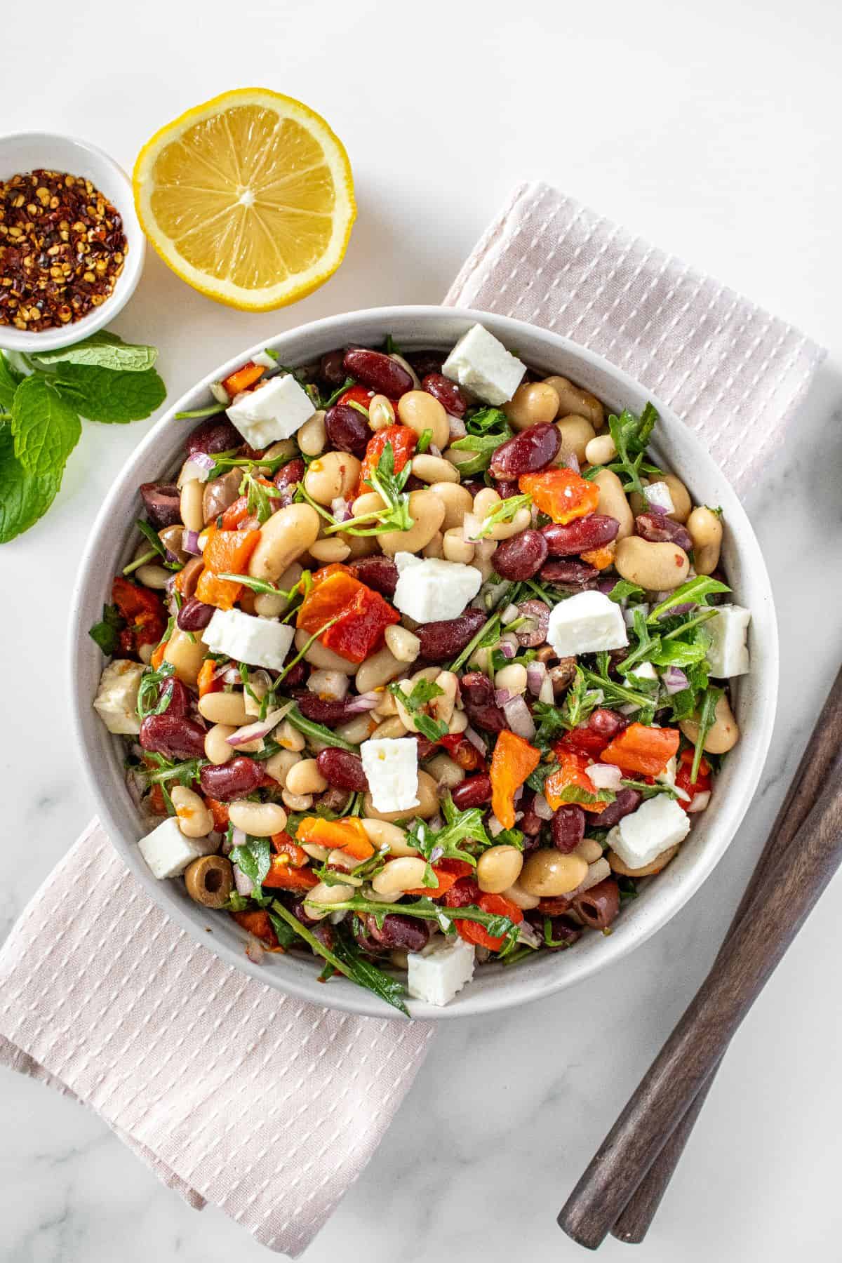 Round white bowl of Mediterranean Bean Salad, on a pink towel, with some garnishes around the edge.