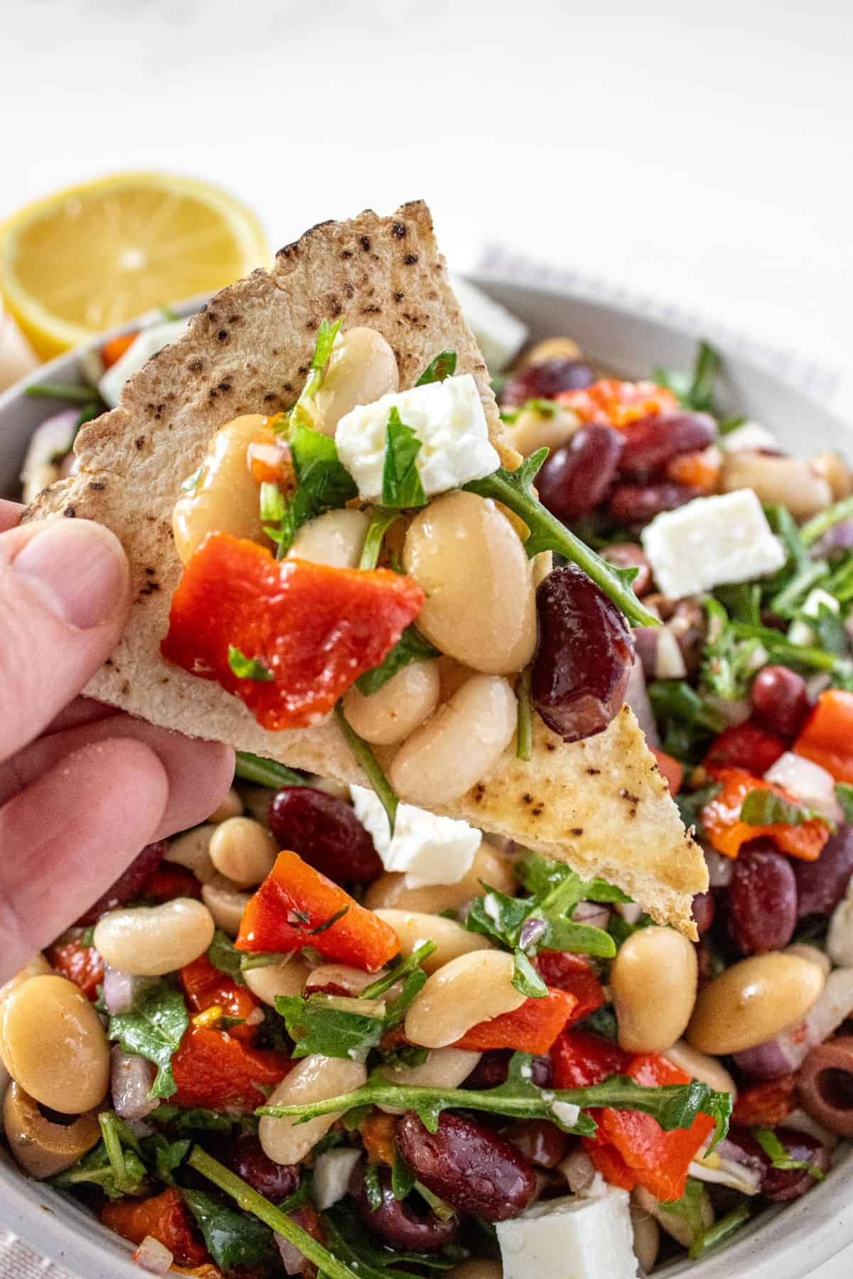 A hand holding a piece of pita bread, with some Mediterranean Bean Salad on top.