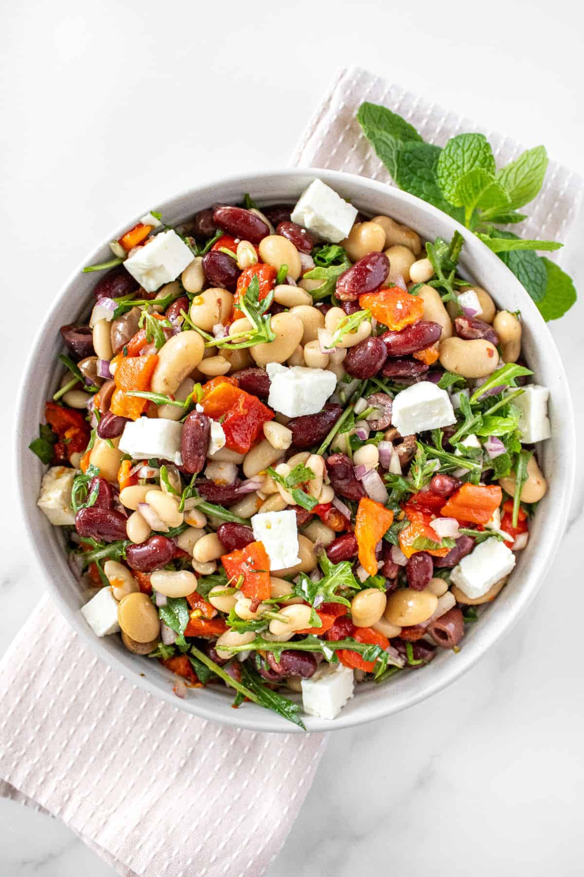 Round white bowl of Mediterranean Bean Salad, on a pink towel, with some garnishes around the edge.