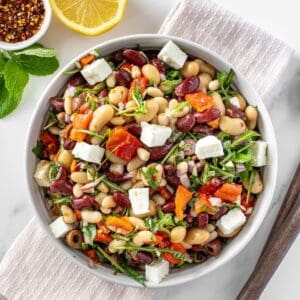Round white bowl of Mediterranean Bean Salad, on a pink towel, with some garnishes around the edge.