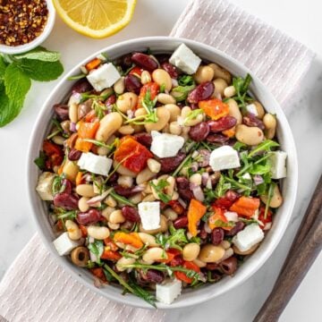 Round white bowl of Mediterranean Bean Salad, on a pink towel, with some garnishes around the edge.