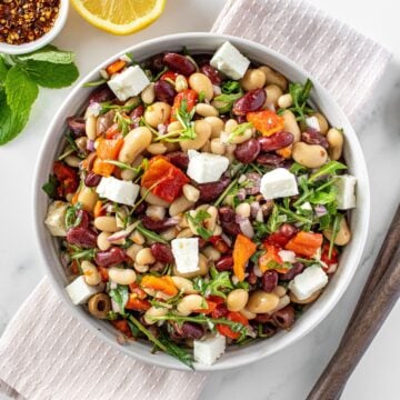 Round white bowl of Mediterranean Bean Salad, on a pink towel, with some garnishes around the edge.