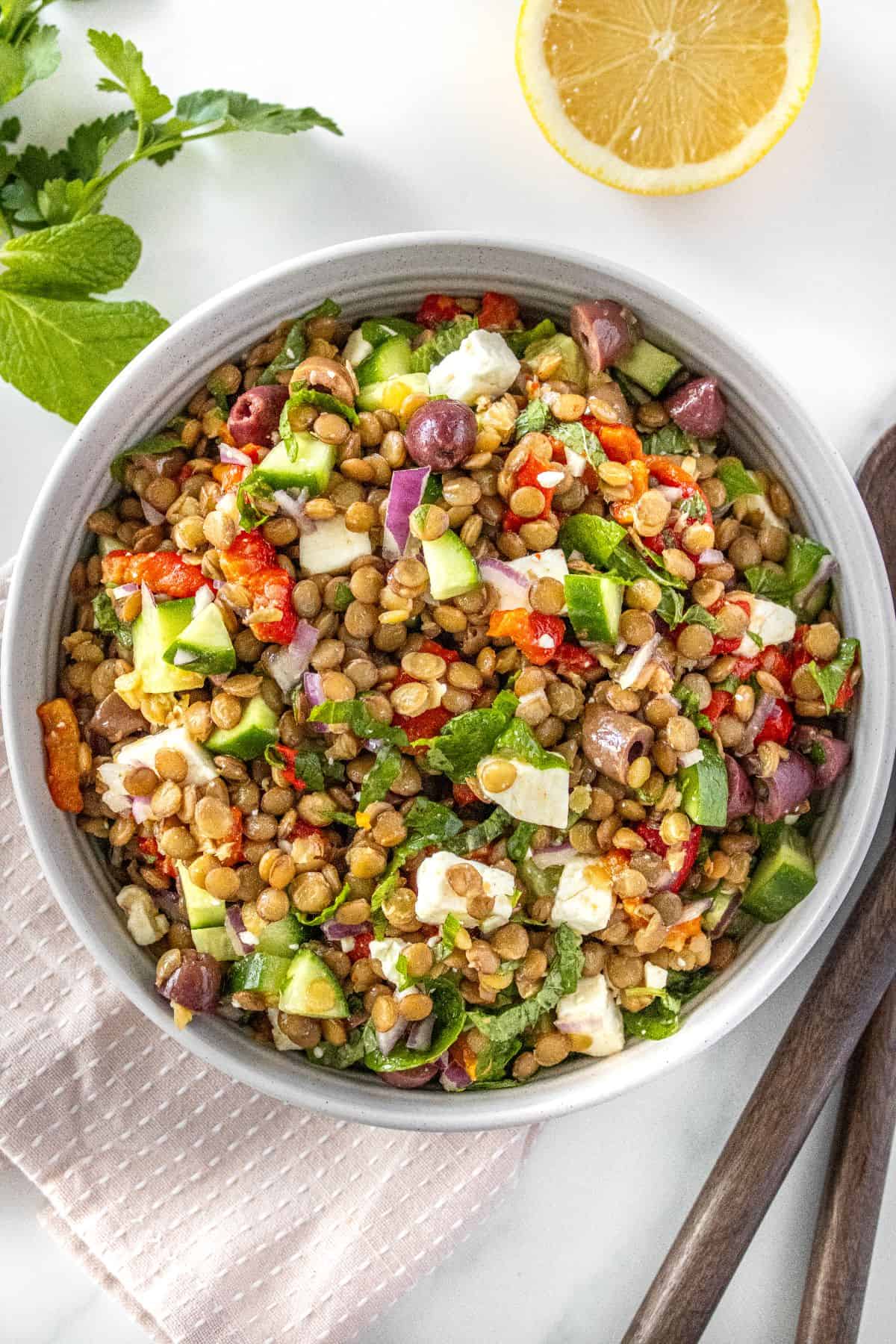 Round bowl of Mediterranean Lentil Salad, sitting on pink cloth, with some herbs, lemon and serving spoons around the bowl.