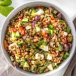 Round bowl of Mediterranean Lentil Salad, sitting on pink cloth, with some herbs and serving spoons around the bowl.