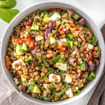 Round bowl of Mediterranean Lentil Salad, sitting on pink cloth, with some herbs and serving spoons around the bowl.