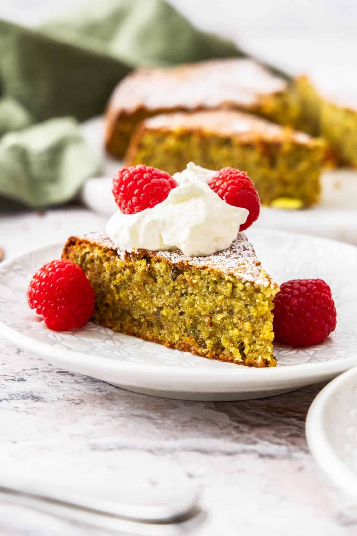 Piece of Pistachio Cake on a small white plate, with the cake topped with whipped cream and fresh raspberries.