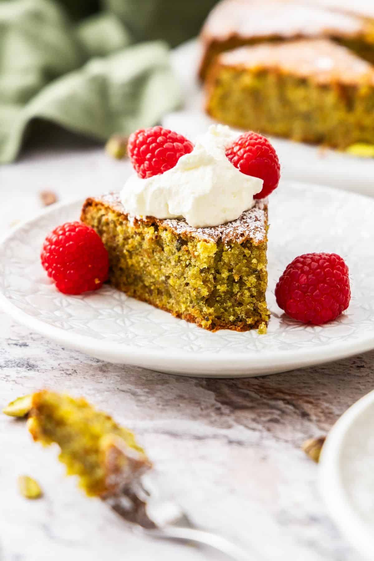 Piece of Pistachio Cake on a small bite missing, with the cake topped with whipped cream and fresh raspberries.
