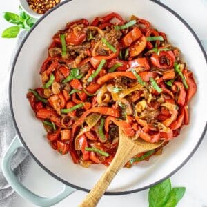 Sautéed Peppers and Onions in a large ceramic dish with a wooden spoon, and some basil leaves and chilli around the edge.