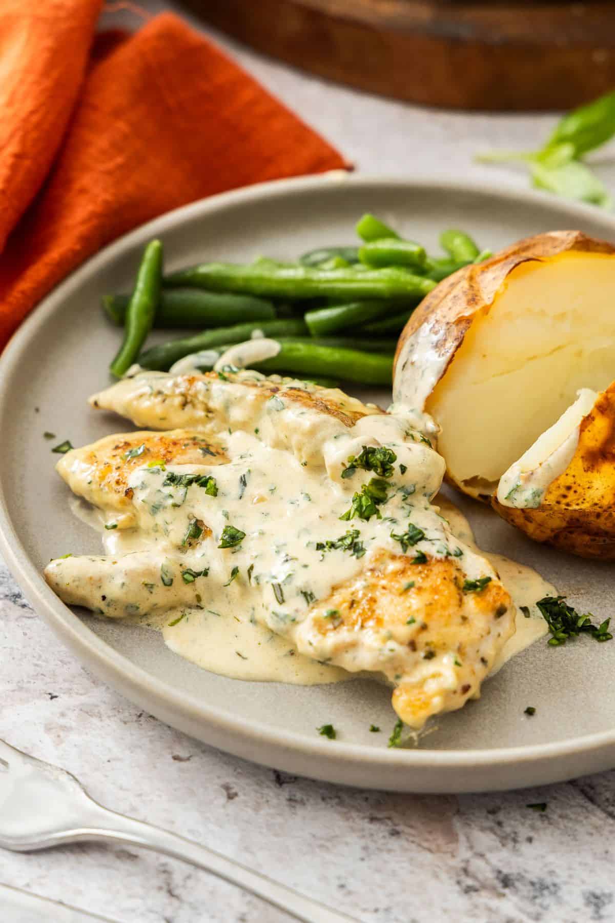 Round plate with serve of Creamy Herb Chicken with a baked potato and some green beans on the side.