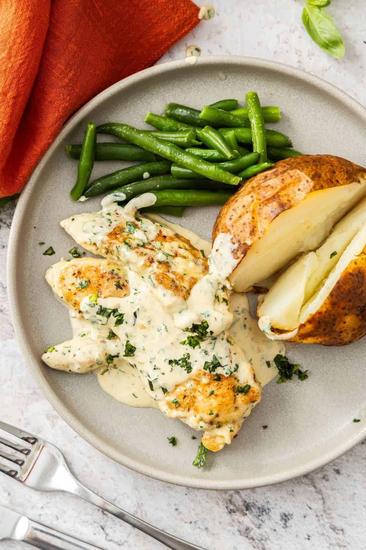 Round plate with serve of Creamy Herb Chicken with a baked potato and some green beans on the side.