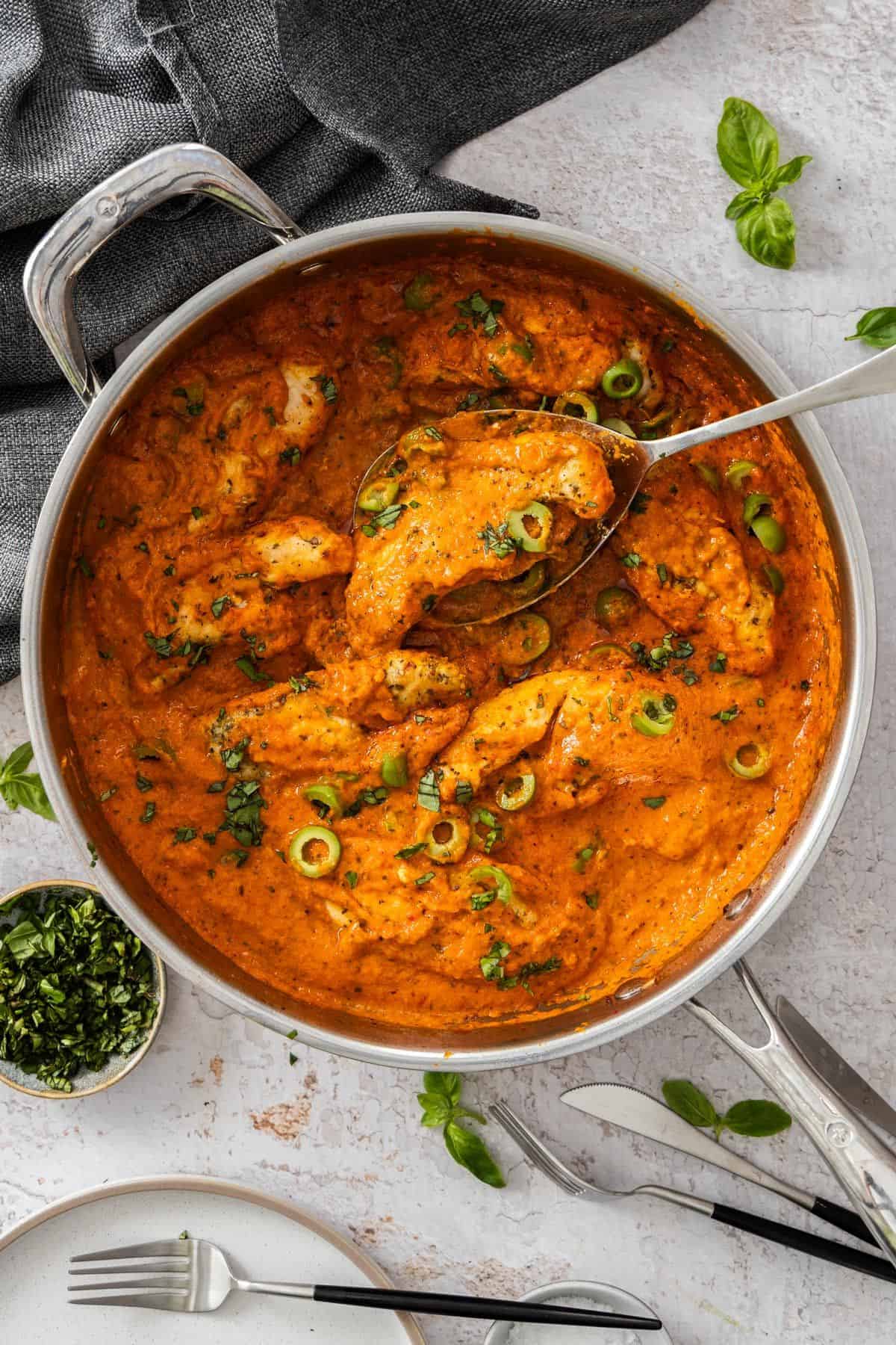 Creamy Red Pepper Chicken in a large frying pan, with a spoon lifting up a piece of chicken.
