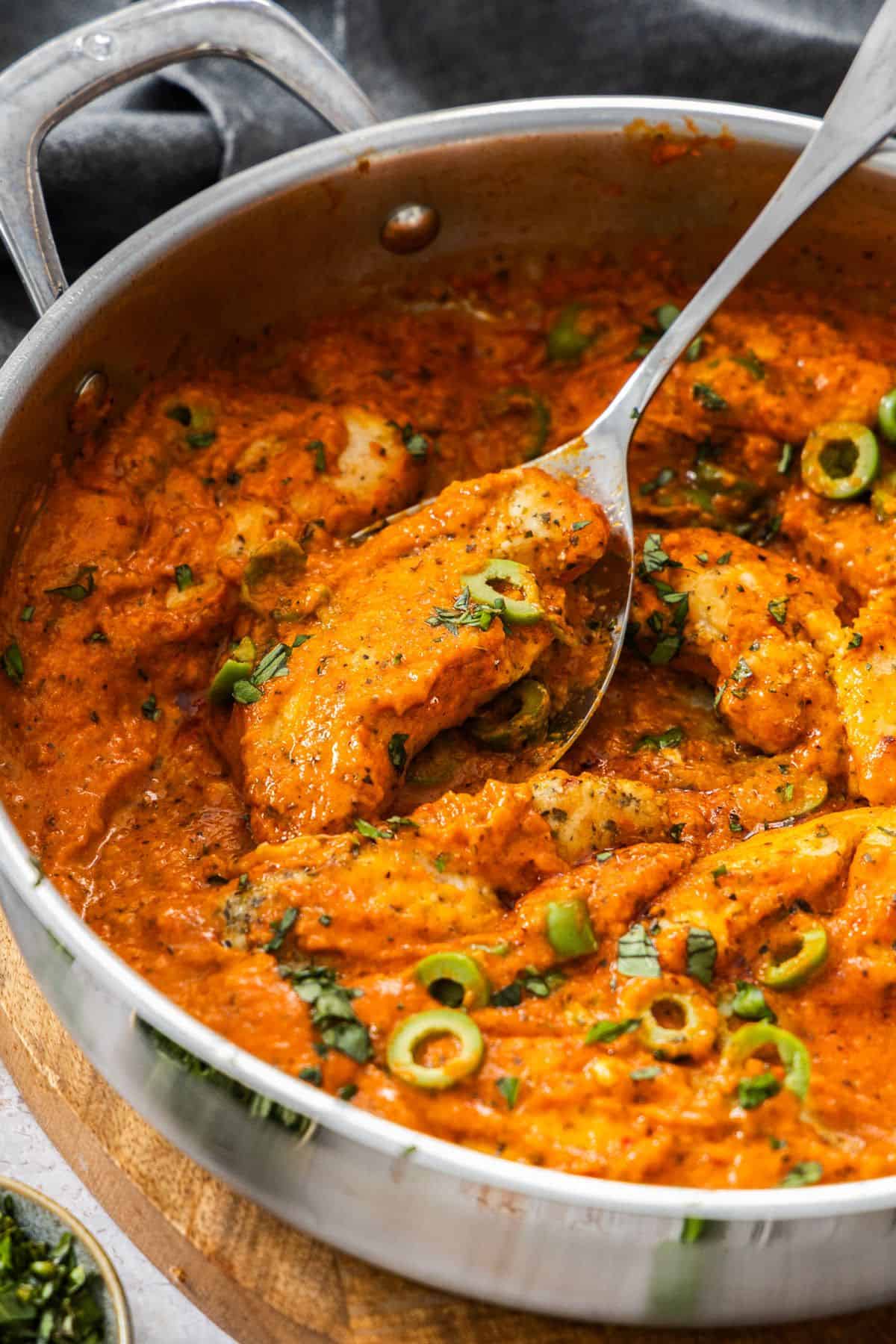Creamy Red Pepper Chicken in a large frying pan, with a spoon lifting up a piece of chicken.