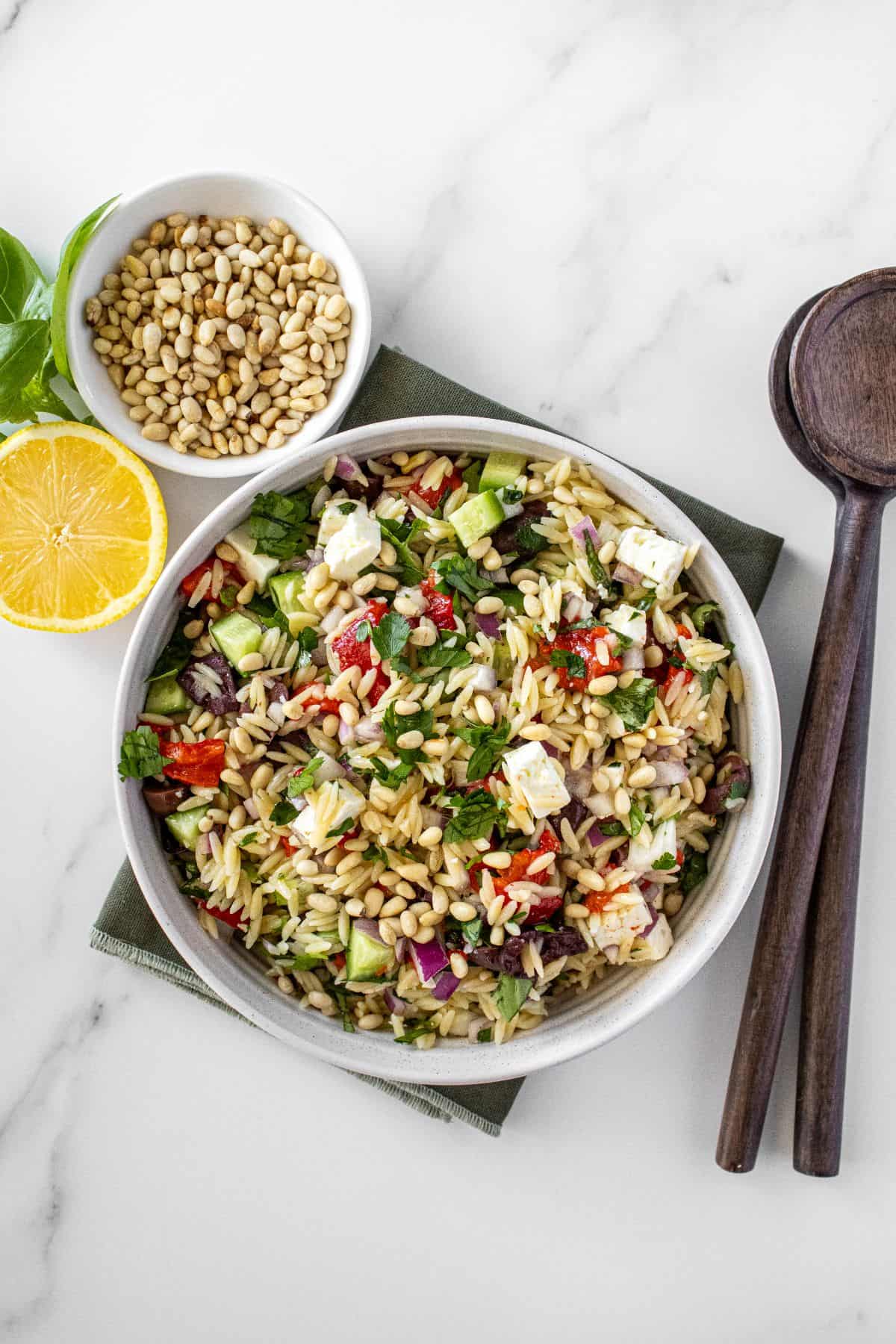 Round white bowl of Mediterranean Orzo Salad, on a green towel, with some garnishes around the edge.