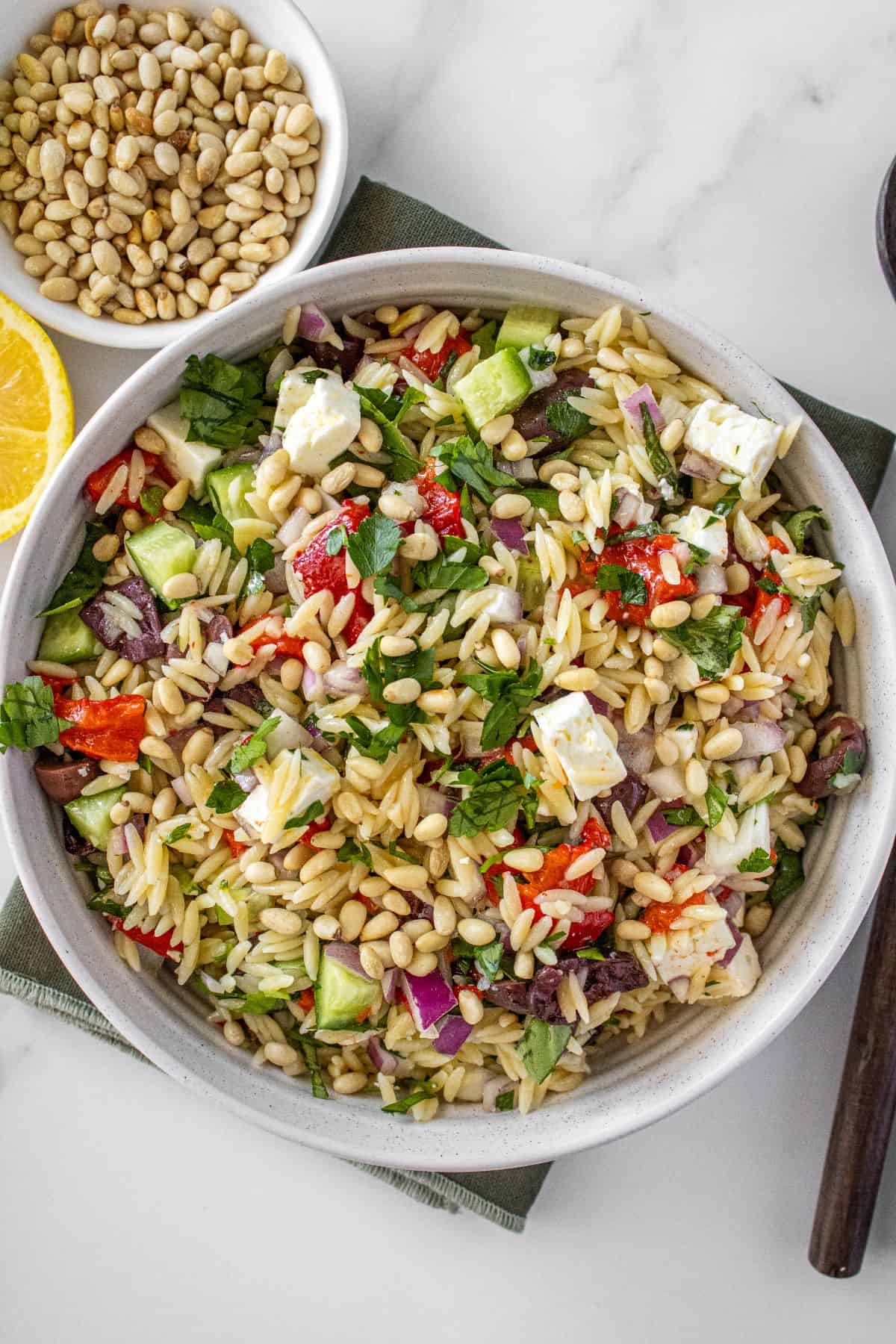Round white bowl of Mediterranean Orzo Salad, on a green towel, with some garnishes around the edge.