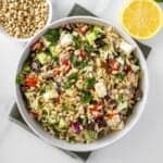 Round white bowl of Mediterranean Orzo Salad, on a green towel, with some garnishes around the edge.