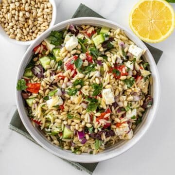 Round white bowl of Mediterranean Orzo Salad, on a green towel, with some garnishes around the edge.