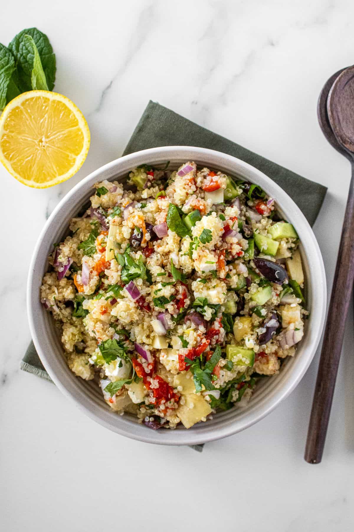 Round white bowl of Mediterranean Quinoa Salad, on a green towel, with some garnishes around the edge.
