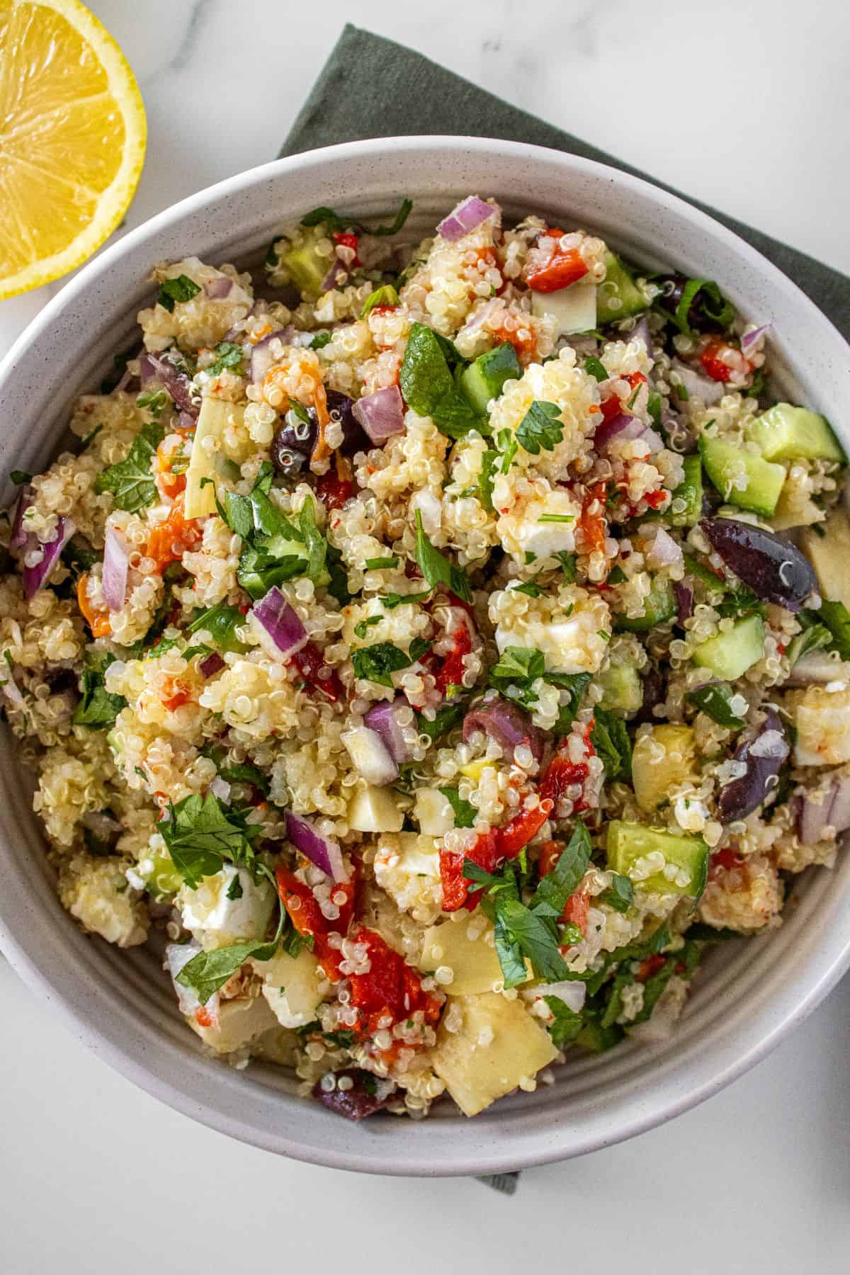 Round white bowl of Mediterranean Quinoa Salad, on a green towel, with some garnishes around the edge.