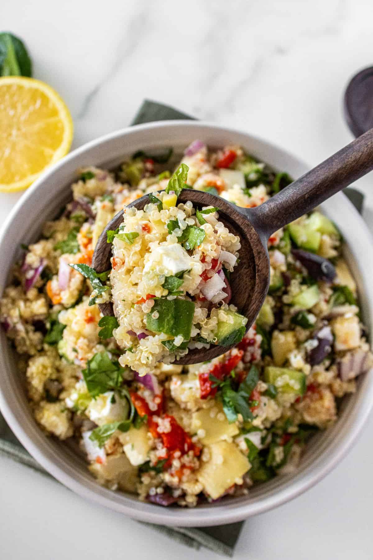 Round white bowl of Mediterranean Quinoa Salad, on a green towel, with a spoon lifting up a scoop of salad.