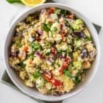 Round white bowl of Mediterranean Quinoa Salad, on a green towel, with some garnishes around the edge.