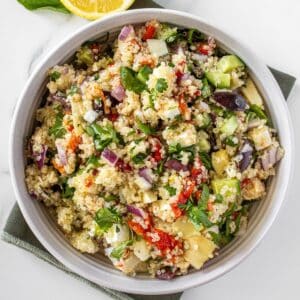 Round white bowl of Mediterranean Quinoa Salad, on a green towel, with some garnishes around the edge.