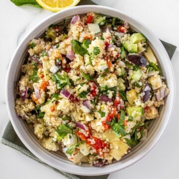 Round white bowl of Mediterranean Quinoa Salad, on a green towel, with some garnishes around the edge.